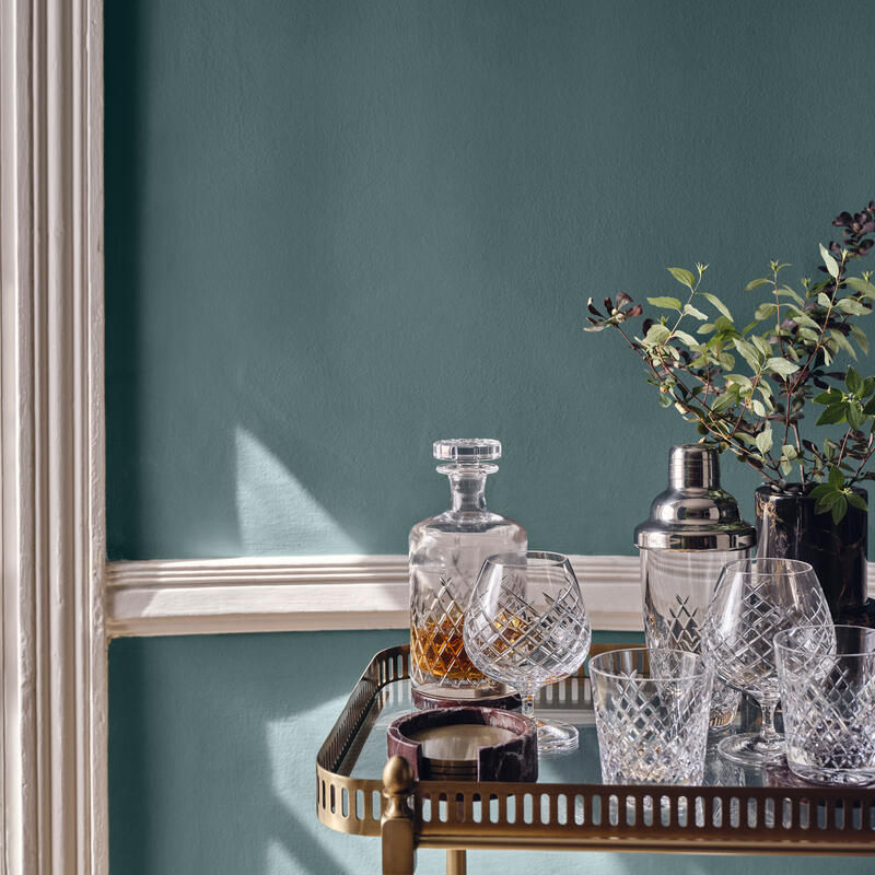 Crystal decanter and glasses sit on a brass bar cart, with a cocktail shaker and a vase of greenery against a teal wall with white trim.