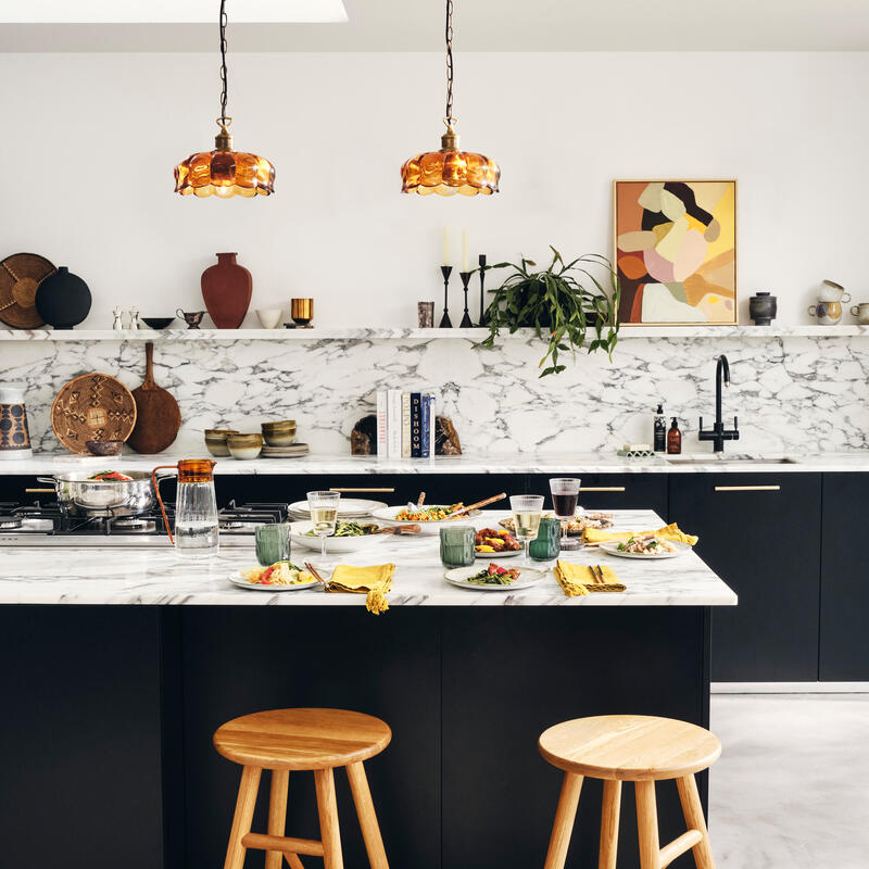 Wooden stools sit by a marble-countered kitchen island set with food and plates, under amber pendant lights; shelves with decor and plants adorn the white, marble-accented wall behind.