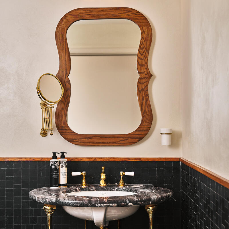 A black marble sink with gold fixtures holds soap bottles, under a large wooden-framed mirror, set in a bathroom with dark tiled walls and a wall lamp above.