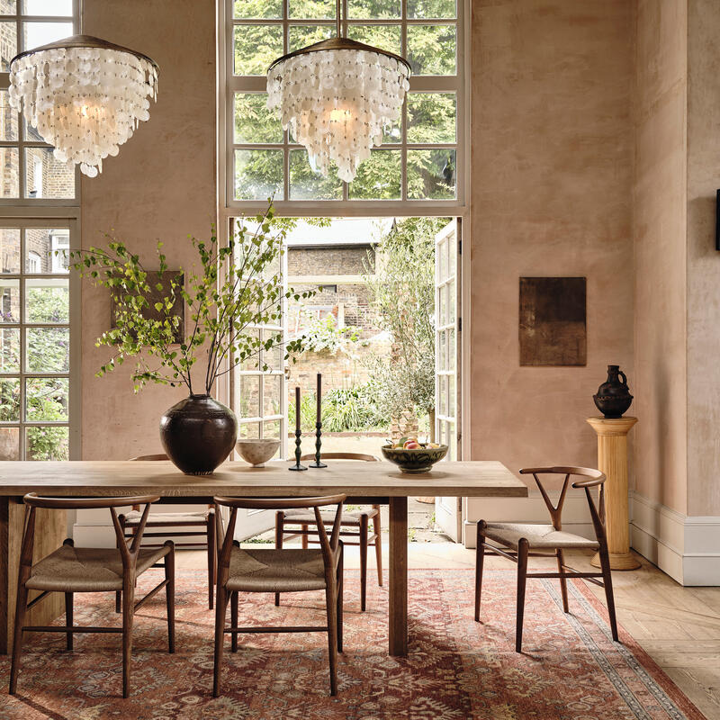 Dining table with chairs arranged around it, decorated with a large vase holding green foliage and candles, situated in a bright room with large windows and chandeliers, overlooking a garden.