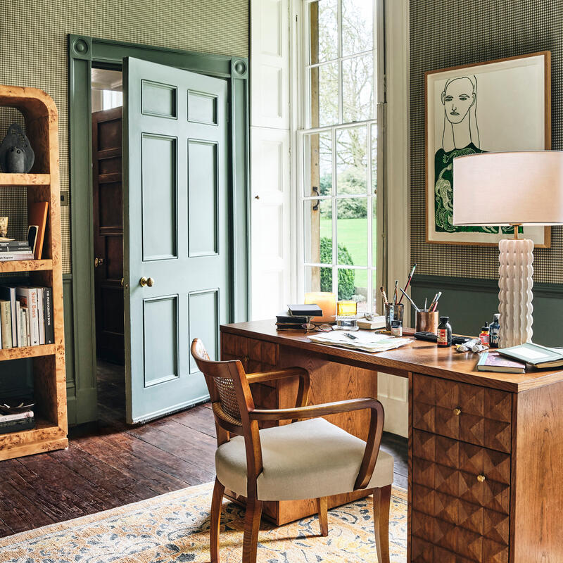 Wooden desk with a chair covered in papers and office supplies, situated in a well-lit study room featuring a patterned rug, bookshelf, closed door, large window, and framed art on the wall.