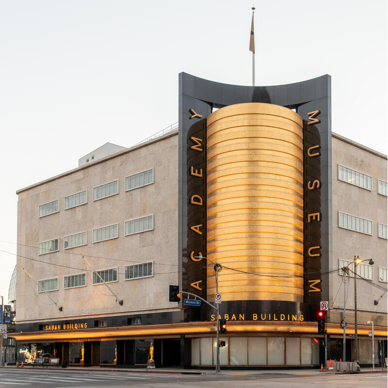 A multi-story beige building with horizontal windows displays vertical "ACADEMY MUSEUM" text beside a golden cylindrical structure labeled "SABAN BUILDING" situated at an urban street corner with cars passing.