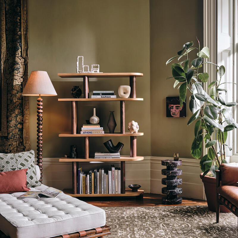 A mid-century modern living room showcasing a cushioned bench, a wooden bookshelf with various books and decorative items, a floor lamp, a large leafy plant, and intricate patterned carpet.