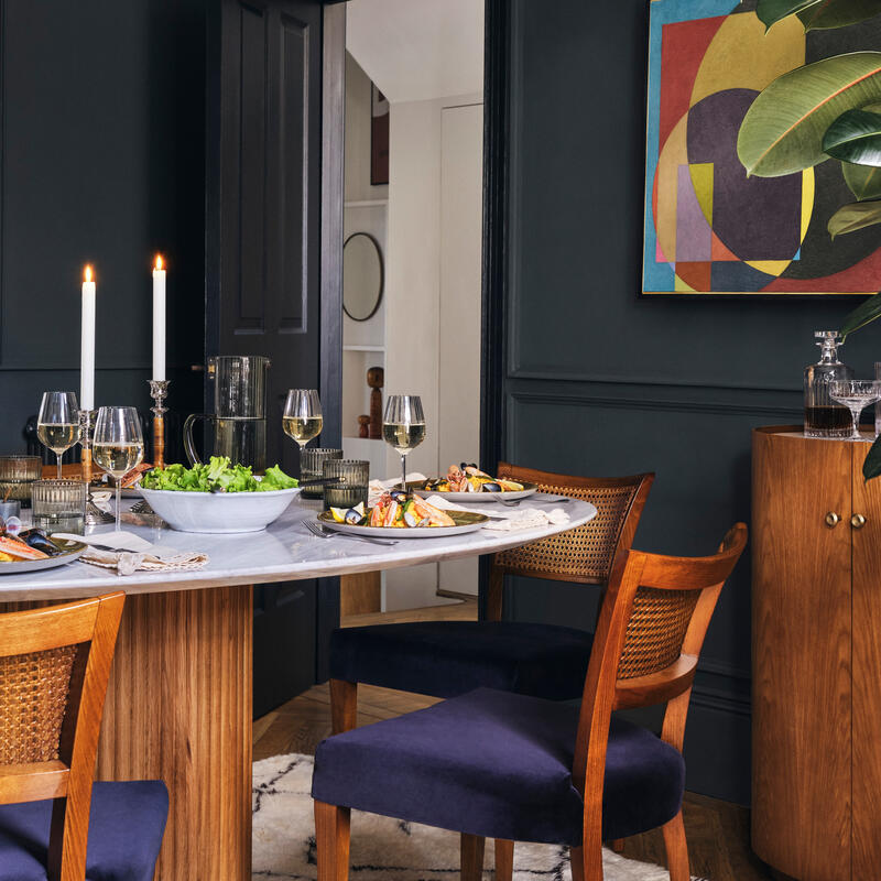 A round marble-topped dining table set for a meal with wooden chairs, two lit candles, and wine glasses; the elegant room features dark walls, modern artwork, and potted plants.