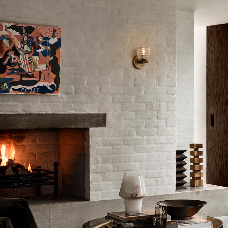 Modern beige sofa with cushions sits on a patterned rug in a cozy living room, featuring a lit fireplace, a black coffee table with decor, armchair, lamp, and abstract wall art.