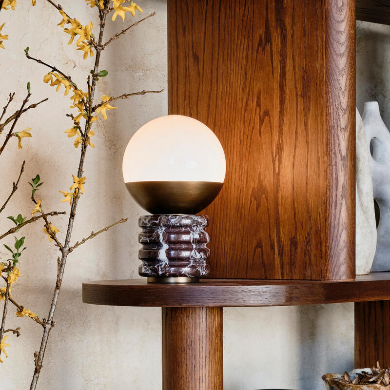 Ceramic vase with yellow flowers and green leaves sits on a marble table beside a lit candle; next to it, a wooden shelf with a round lamp, books, and black decorations.