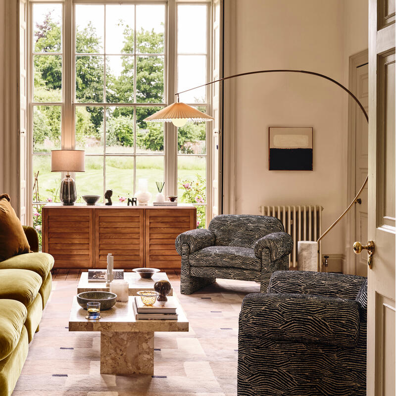 A green plush sofa and two patterned armchairs face a stone coffee table in a well-lit living room with a large window overlooking a garden. A wooden sideboard and potted plant complete the setting.