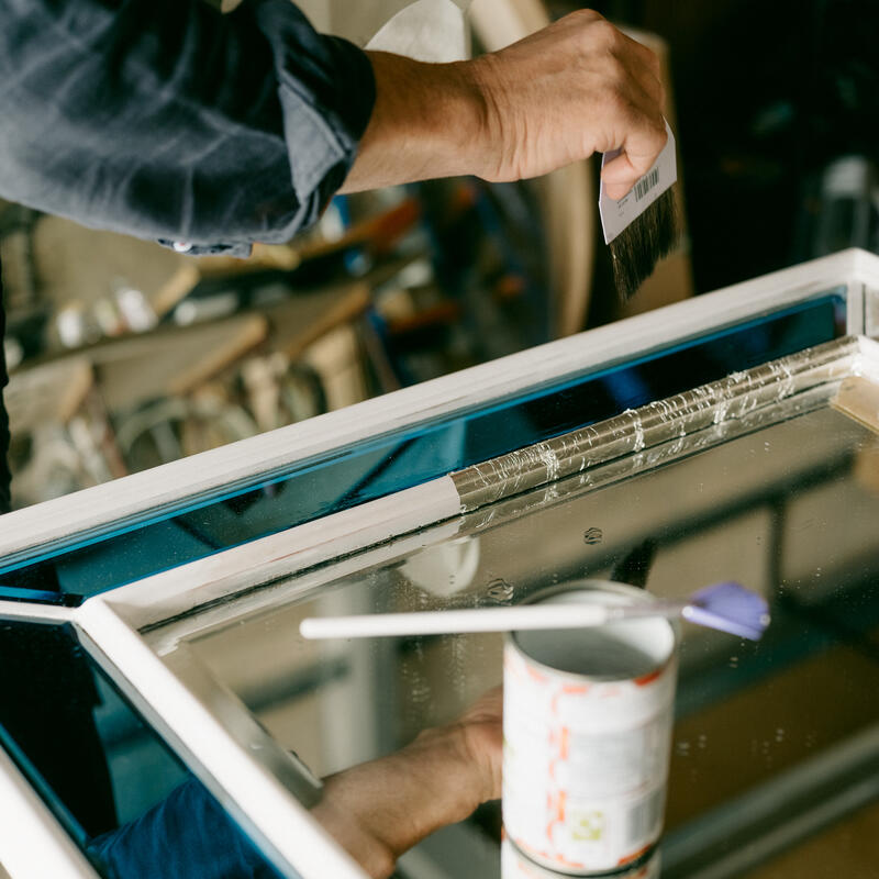 A hand holding a brush applies primer or paint to a window frame in a workshop with various tools and supplies in the background.