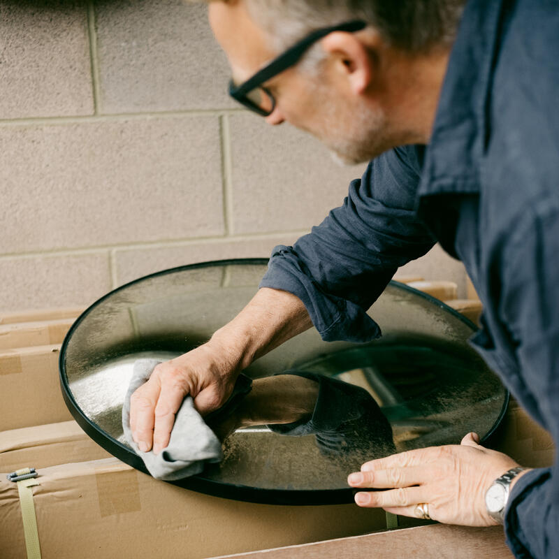 A man with glasses is meticulously cleaning a large, circular glass object with a cloth in a workspace, surrounded by cardboard boxes against a beige cinderblock wall.