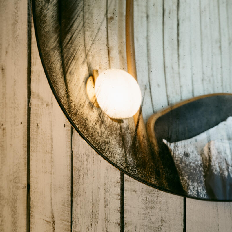 A round mirror reflecting a lit bulb and vertical wooden panels, with visible dust and smudges on the mirror surface, mounted on whitewashed wooden planks.