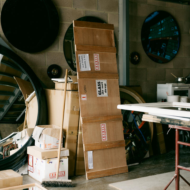 A collection of mirrors and cardboard boxes, some labeled "FRAGILE, GLASS," are stacked against a concrete wall in a cluttered warehouse. A broom leans against the boxes.