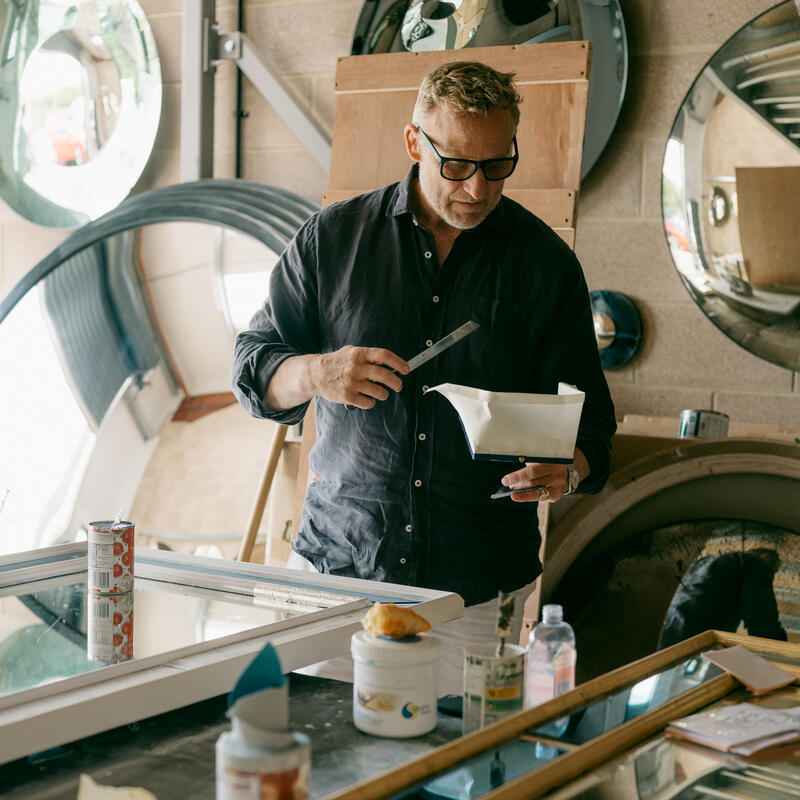 A man in a black shirt and glasses examines a white object with a tool in an industrial workshop, surrounded by mirrors and various containers on a large table.