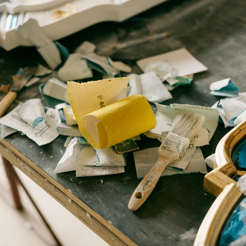 Yellow sandpaper and a paintbrush rest among scattered paper scraps on a workshop table; a partially painted wooden frame lies nearby.