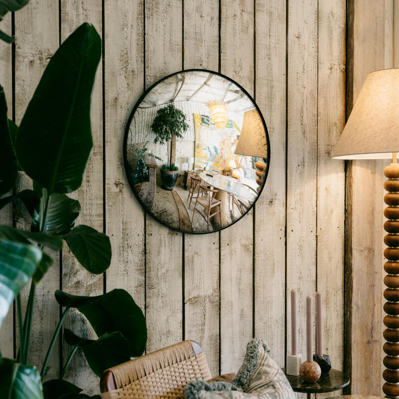 A round mirror reflecting a cozy, plant-filled indoor space hangs on a rustic wooden wall. Nearby, a wicker chair with patterned pillows and a side table with candles complete the inviting scene.