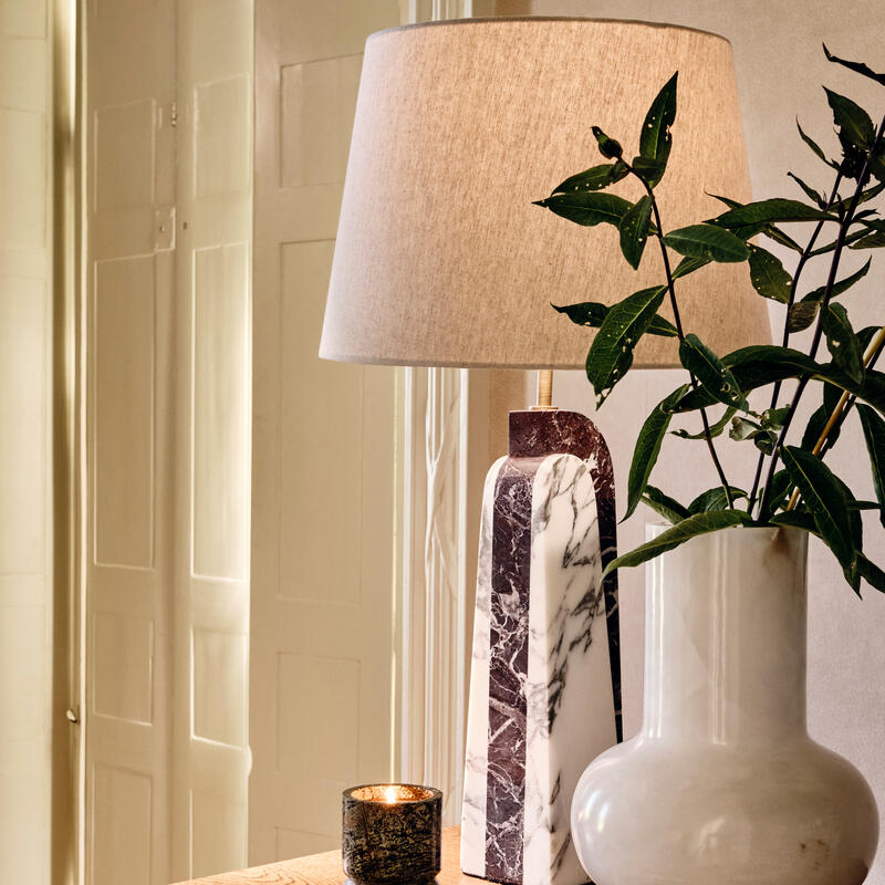 A table lamp with a beige shade and marble base sits beside a vase with green leaves and a candle on a wooden dresser in a softly lit room.