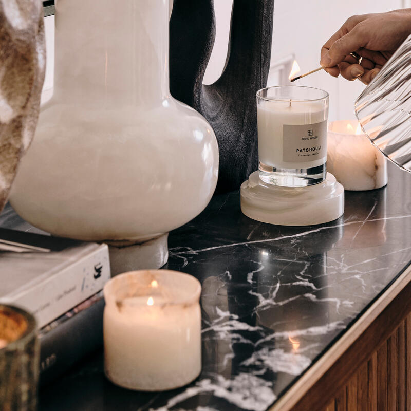 A hand lights a "Patchouli" candle with a match on a marble surface, surrounded by decorative objects, including white and brown vases and other candles.