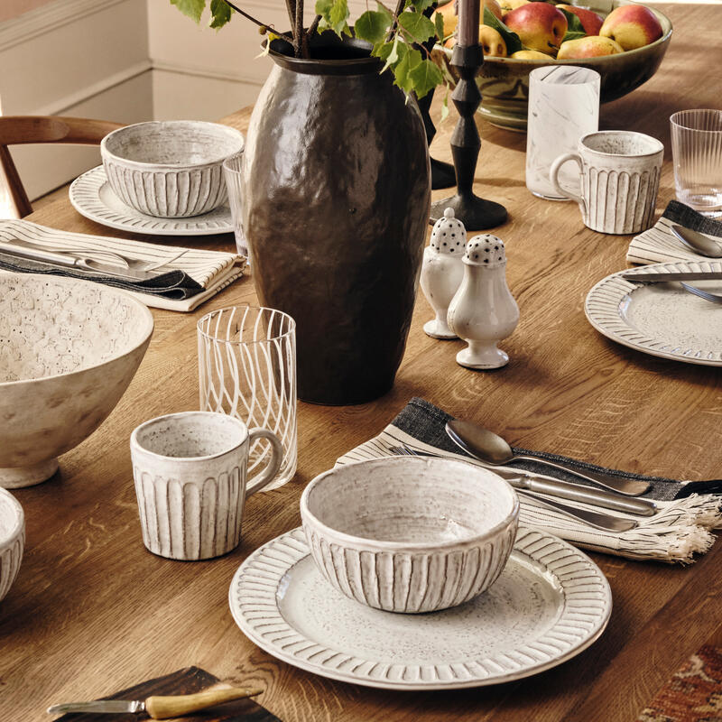 Ceramic bowls, plates, mugs, and glasses, along with cutlery on napkins, are arranged on a wooden dining table. A large dark vase with green plant, salt and pepper shakers, and a fruit bowl complete the setting.