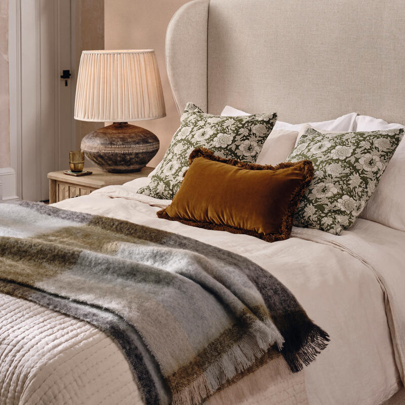 A beige bed with floral pillows and a brown decorative cushion is neatly made in a cozy, warmly-lit room with a wooden nightstand, a table lamp, and an arched doorway.