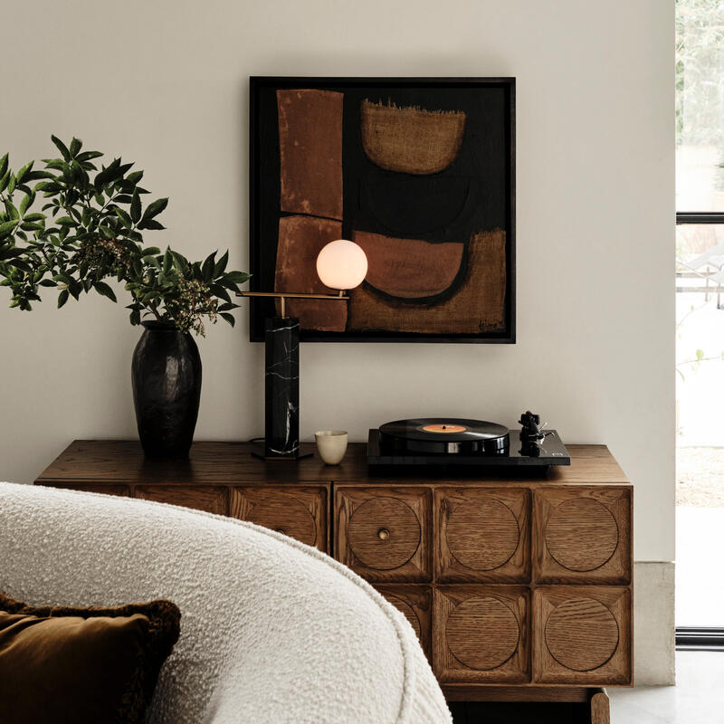 A wooden sideboard holds a black vase with green foliage, a black lamp, a small cup, and a turntable in a living room with modern art and a white curved sofa.