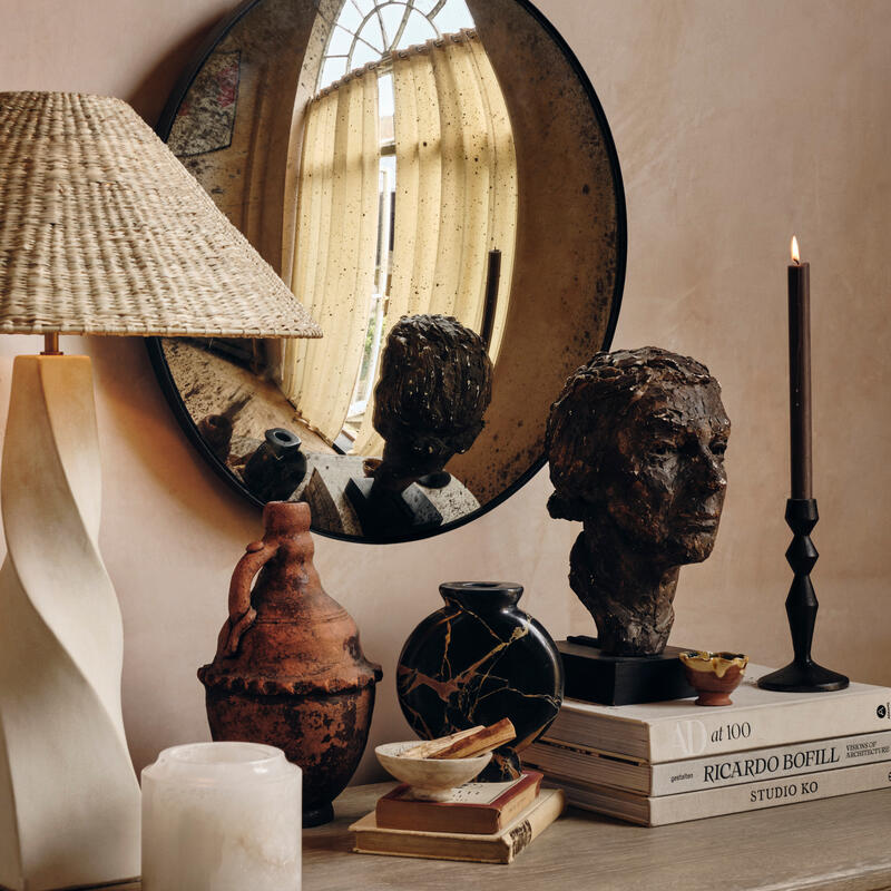 Various decorative objects rest on a wooden dresser, including a lamp, sculptures, vases, a candle, and books ("AD at 100", "Ricardo Bofill", "Studio KO"). A round mirror reflects a window.