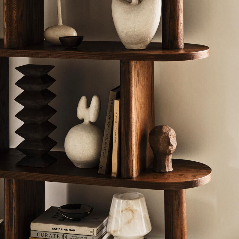 Various abstract sculptures, bowls, and books are arranged on a wooden shelf in a minimalistic, neutral-toned room. Books visible are "Portraits of Home," "CURATE," and "EAT DRINK NAP."