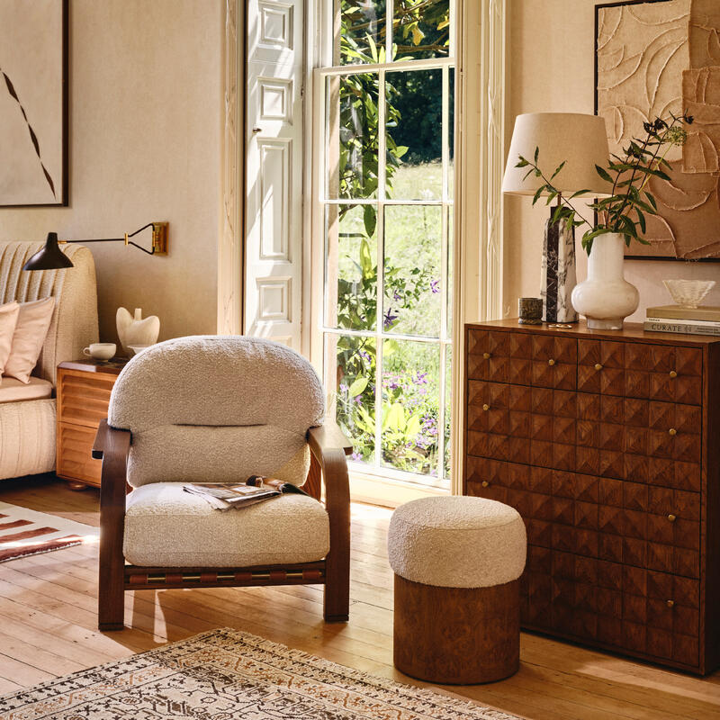 A cozy armchair and a small cushioned stool rest on a patterned rug in a sunlit room, with a leafy garden visible through tall, open French windows.