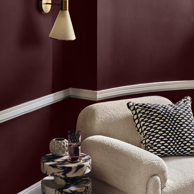 Twin-cone wall lamp illuminates a plush beige armchair with a patterned cushion; a round marble side table holds a textured vase and a faceted glass against burgundy walls with white trim.