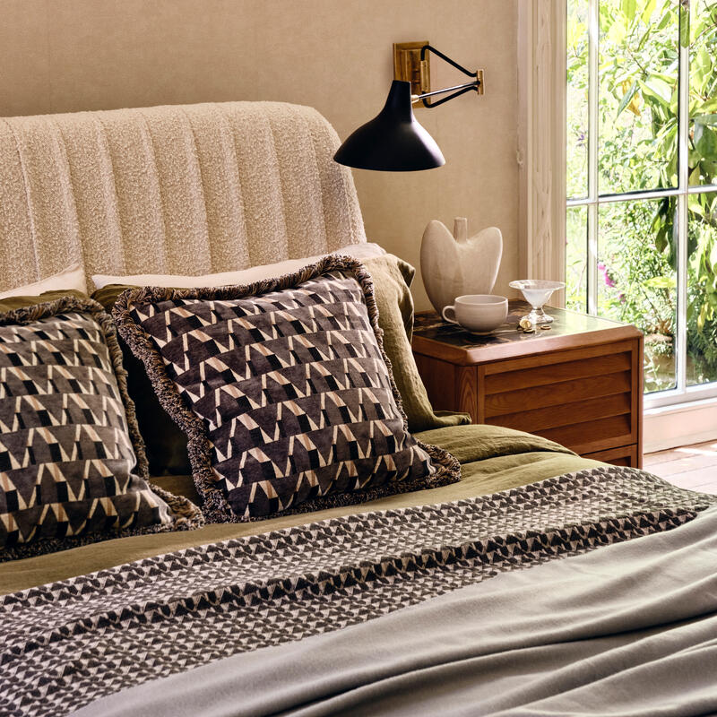 Bed arranged with multicolored geometric-patterned cushions, green bedspread, and gray blanket; accompanied by a wooden nightstand with a ceramic vase and cup beneath a black wall lamp. Sunlit window and leafy outdoor view.