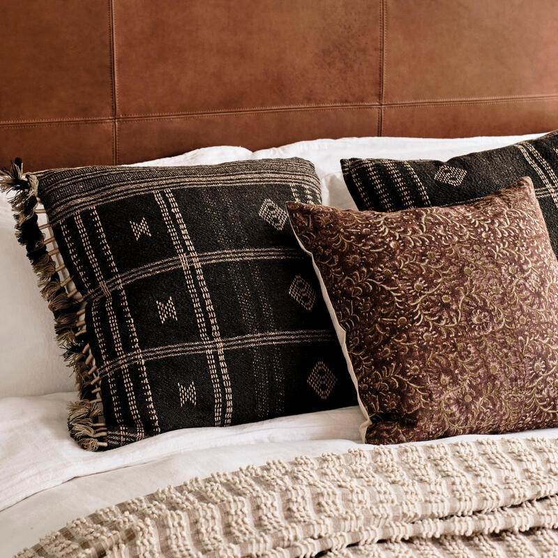 Decorative pillows, two patterned and one embroidered, rest on a white bed with a brown leather headboard, creating a cozy, sophisticated bedroom setting. A textured beige throw is laid across the bed.