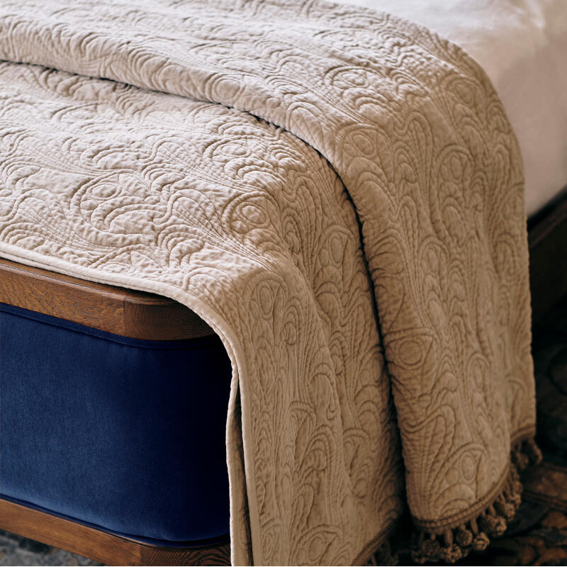 A beige, patterned throw blanket with tassels drapes over the edge of a navy-blue upholstered bed with wooden frame, set atop a floral-patterned rug.