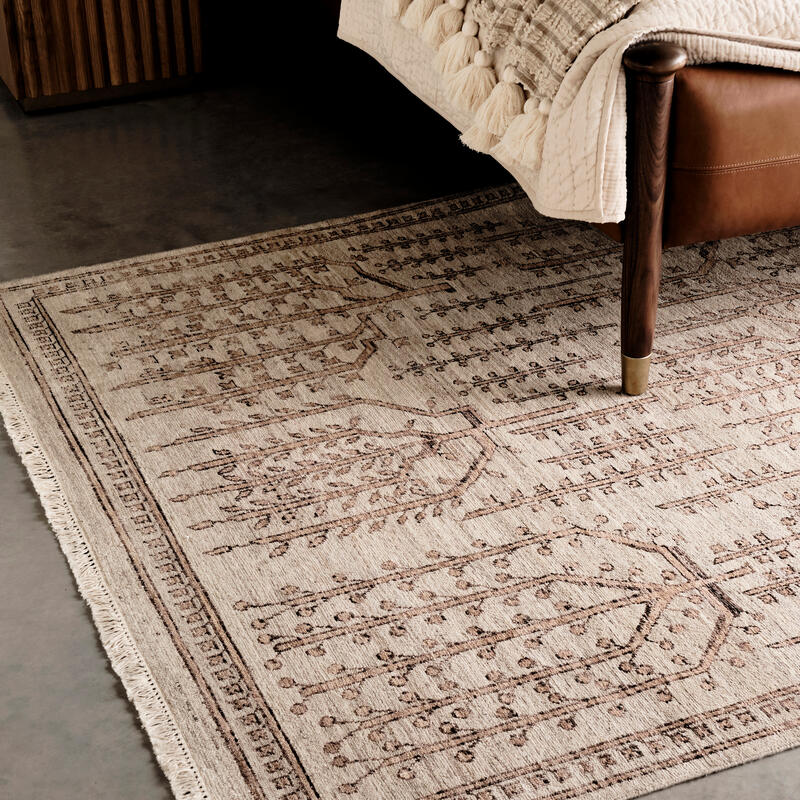 A detailed, patterned rug lies on a polished concrete floor beside a bed with a quilted blanket and tassels. A wooden nightstand stands in the background.