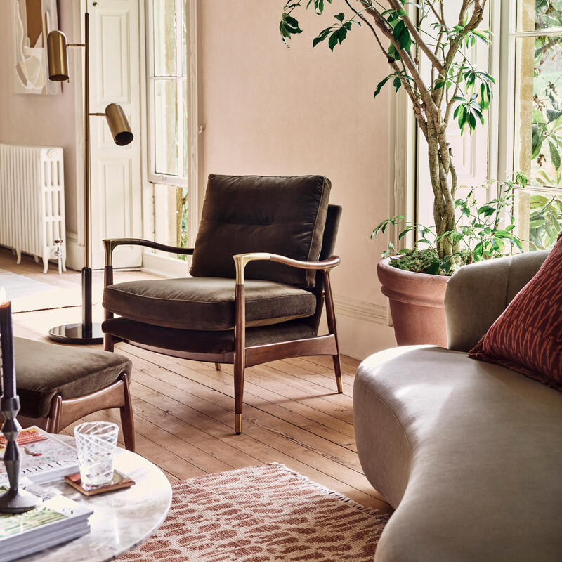 A cushioned armchair rests near a large potted plant beside a tall window in a bright, elegantly furnished living room with an ornate floor lamp, a rug, and a round coffee table.