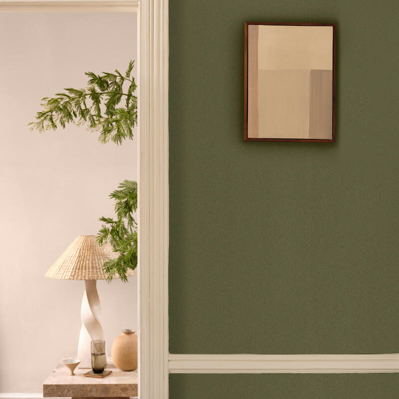 A framed abstract painting hangs on a green wall with a white doorframe. Through the doorway, a decorative table with a woven lampshade, a plant, and ceramic items is visible.