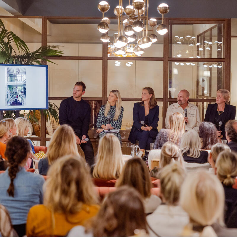 A panel of six people holding microphones speaks to a seated audience in a modern room with a television and plants, while contemporary light fixtures hang overhead.