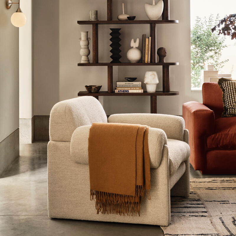 A beige armchair with an orange throw blanket sits in a modern living room; behind it, a wooden shelf displays various ceramics and books, with natural light entering from a window.