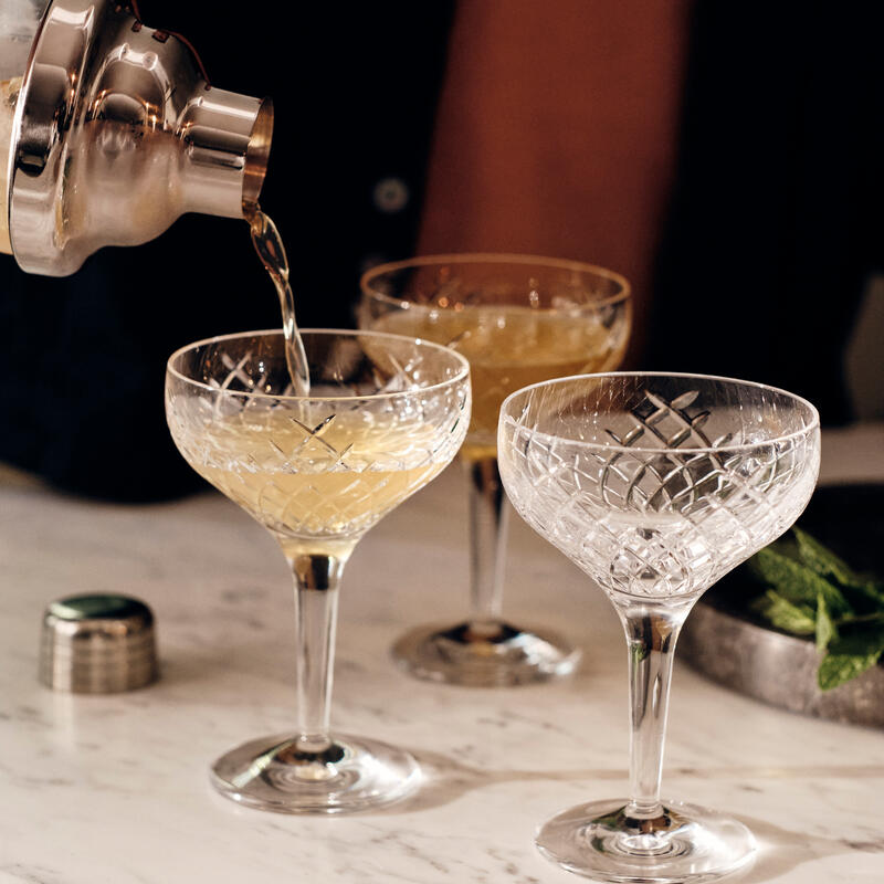 Cocktail shaker pouring drink into one of three etched coupe glasses on marble countertop near a small metal cap and a mint-filled bowl.