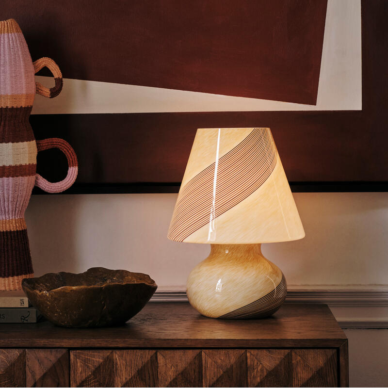 A sculptural table lamp with a swirling pattern is lit and placed on a wooden sideboard next to a textured bowl and a decorative object. The background features abstract geometric artwork.