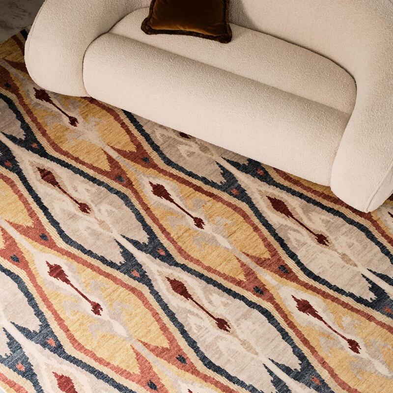 Beige sofa with a brown pillow sits on a patterned rug featuring yellow, red, and dark blue diamond shapes and abstract designs in a minimalist living room.