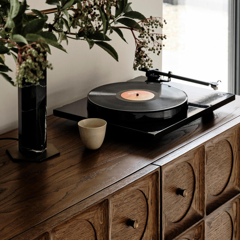 A vinyl record player sits on a wooden cabinet, accompanied by a ceramic cup and a vase with green foliage. Natural light streams in through a nearby window.