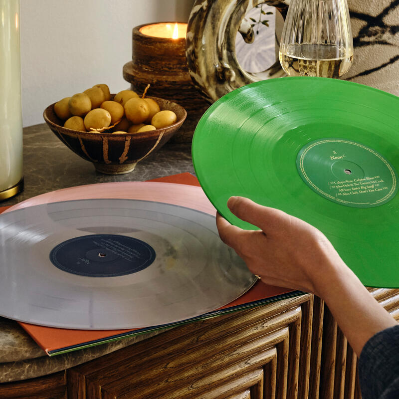 A hand holds a green vinyl record beside a playing grey vinyl record on a marbled table with a fruit bowl, candle, plant, and wine glass in the background.