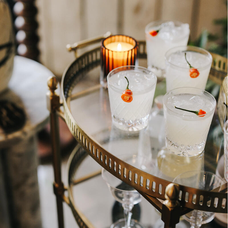 Cocktail glasses with ice and red chili peppers sit on a brass and glass bar cart. A lit candle in a striped holder adds to the upscale, cozy setting indoors.