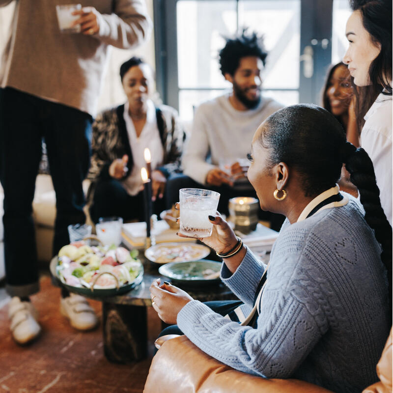 A group of people socializes in a cozy room. One person in the foreground, wearing a blue sweater, sits holding a drink while others gather around a coffee table with food and candles.