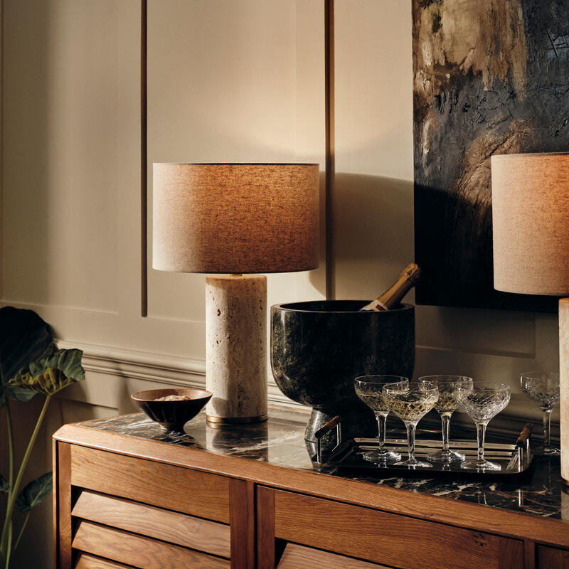 A sideboard holding a lit lamp, a large bowl with a champagne bottle, and a tray of crystal glasses, situated in an elegantly furnished room with beige paneled walls.