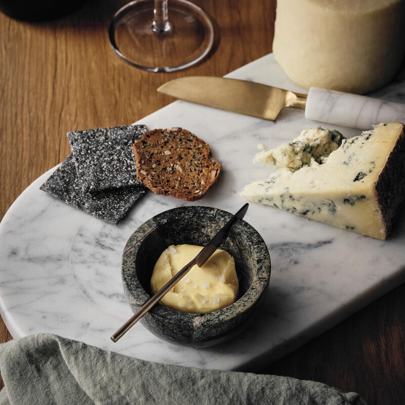 Cheese on a marble board is paired with crackers and butter in a small bowl, next to a wine glass and bottle, all on a wooden table with a green cloth napkin.