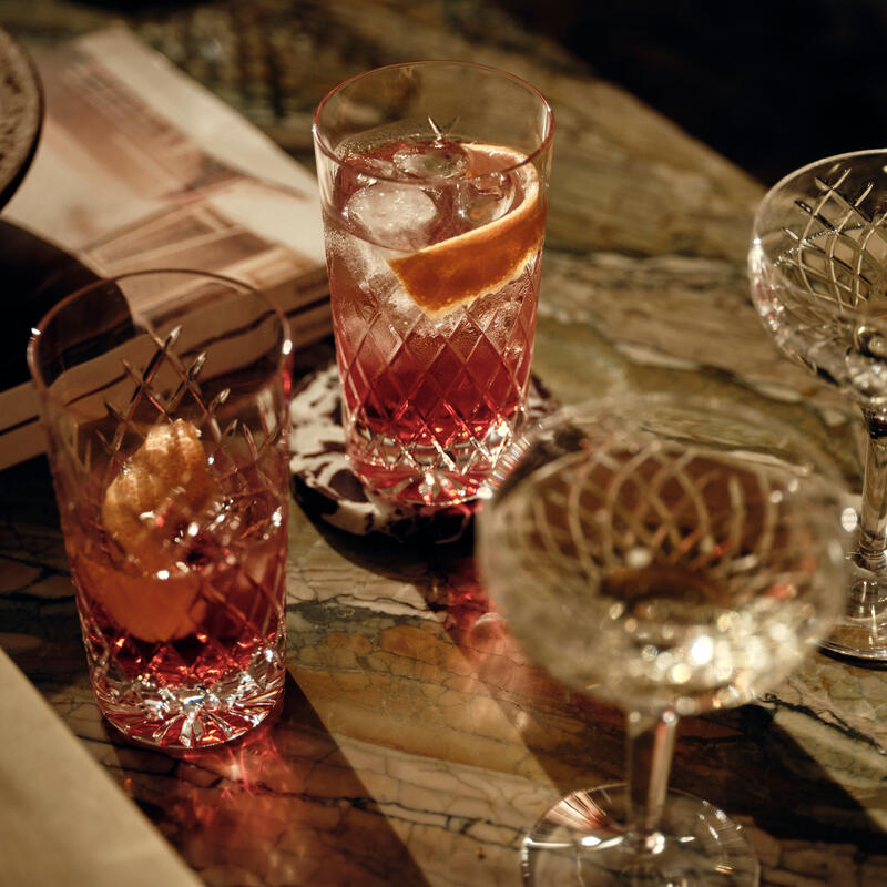 Ornate crystal glasses hold red cocktails with large ice cubes and orange slices; set on a marble table alongside books and a ceramic bowl.