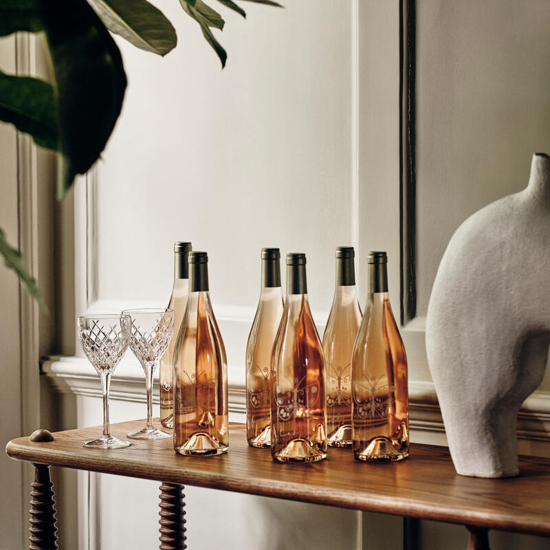 Six rose wine bottles stand on a wooden table next to two crystal wine glasses. The setting is a bright, elegant room with a white wall, greenery, and a sculptural decor piece.