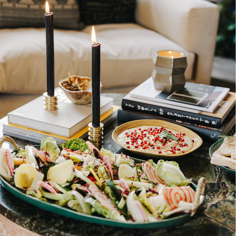 A salad platter with various fresh vegetables on a marble coffee table, accompanied by books, two lit black candles, and a plate of dip garnished with pomegranate seeds in a cozy living room.