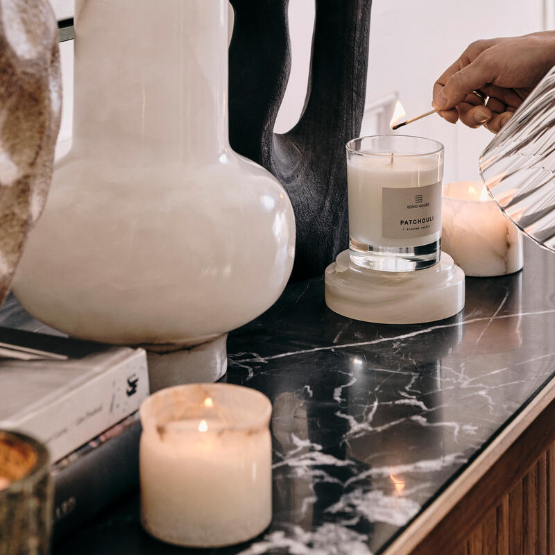 A hand lights a "Patchouli" candle using a matchstick on a marble surface, surrounded by decorative sculptures, vases, and other lit candles.