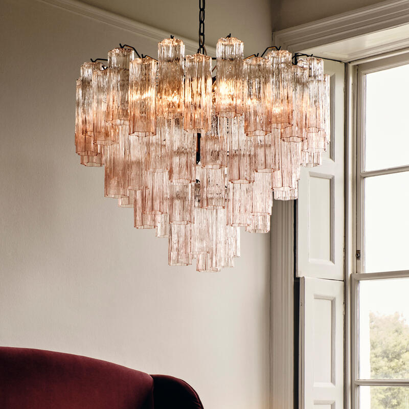 A cascading crystal chandelier hangs above a bed with a deep red upholstered headboard, next to a window with sunlight streaming in and a bedside table holding a lamp and a glass.