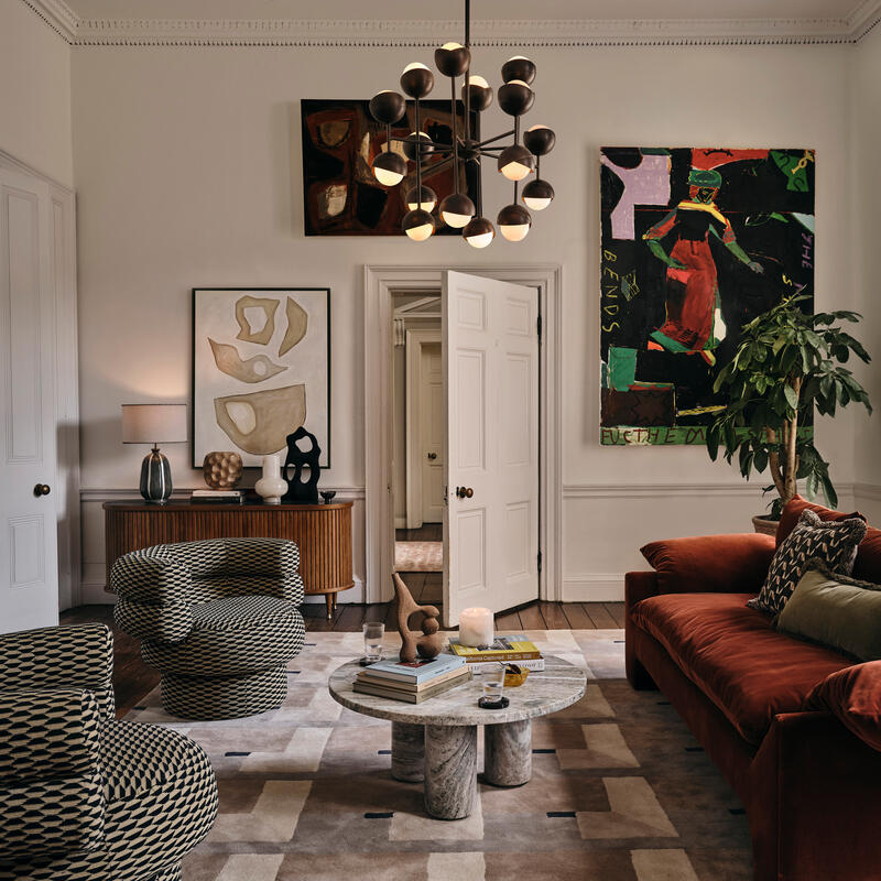 A stylish living room features a rust-colored sofa, patterned armchairs, and a round marble coffee table. Art pieces adorn the walls, and a large window allows natural light in.